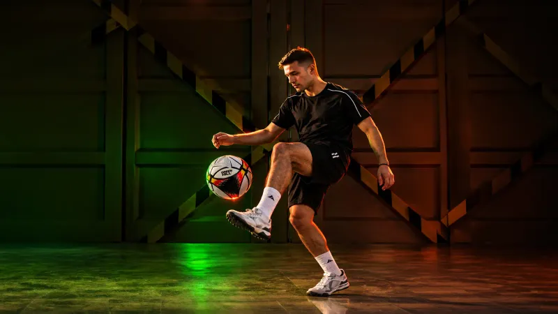 Footballer in a black kit juggling a Green Hill match ball in a dim hall lit in green and amber